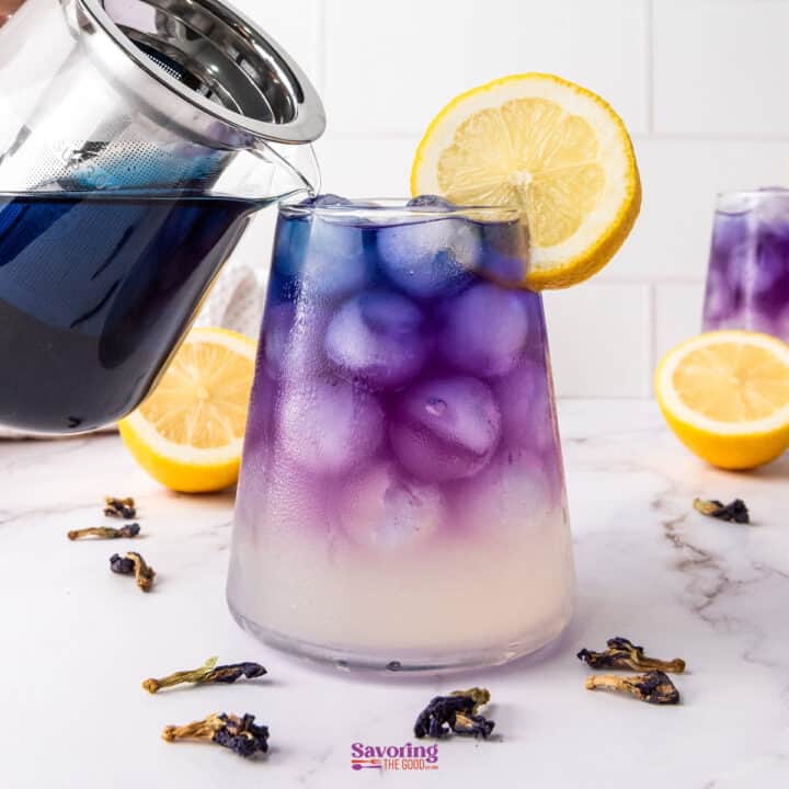 A glass of butterfly pea flower lemonade is being poured, displaying a gradient from blue to purple, garnished with a lemon slice. Lemons and dried flowers are scattered on the white surface.