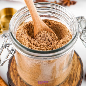 A glass jar filled with ground cinnamon powder, reminiscent of a chai spice mix, with a wooden spoon resting inside. The jar is placed on a wooden coaster.