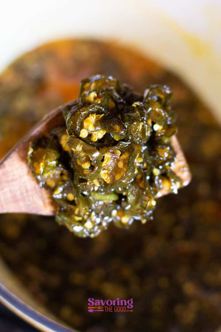 Close-up of candied jalapeños on a wooden spoon, with a pot containing more in the background.