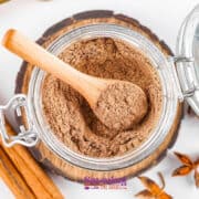 A glass jar brimming with chai spice mix holds a wooden spoon, surrounded by cinnamon sticks and star anise on a charming wooden coaster.