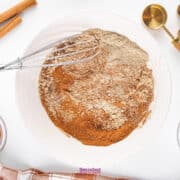 A bowl with mixed dry ingredients, featuring a fragrant Chai Spice Mix, along with a whisk, measuring spoons, cinnamon sticks, and spice bowls on a pristine white surface.