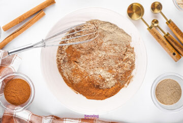A bowl with mixed dry ingredients, featuring a fragrant Chai Spice Mix, along with a whisk, measuring spoons, cinnamon sticks, and spice bowls on a pristine white surface.