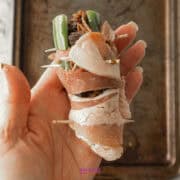 A hand holds a Texas Twinkie, a bacon-wrapped jalapeño stuffed with cheese and vegetables, secured with toothpicks, hovering over a baking tray.