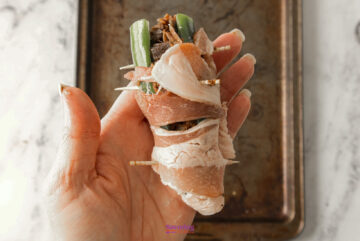 A hand holds a Texas Twinkie, a bacon-wrapped jalapeño stuffed with cheese and vegetables, secured with toothpicks, hovering over a baking tray.