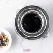 Top view of a teapot filled with vibrant blue liquid, resembling refreshing Butterfly Pea Flower Lemonade, beside a bowl of dried butterfly pea flowers on a white marble surface.