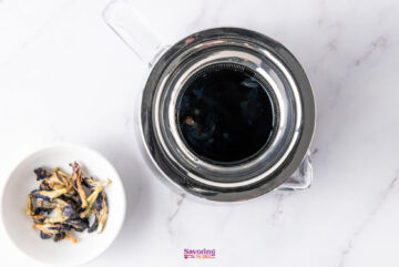Top view of a teapot filled with vibrant blue liquid, resembling refreshing Butterfly Pea Flower Lemonade, beside a bowl of dried butterfly pea flowers on a white marble surface.