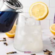 A person pours a dark liquid from a glass teapot into a glass with ice and a lemon slice, crafting a refreshing concoction. In the background, a similar glass contains Butterfly Pea Flower Lemonade, its purple hue complementing the scattered lemon halves and blue petals.