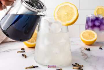 A person pours a dark liquid from a glass teapot into a glass with ice and a lemon slice, crafting a refreshing concoction. In the background, a similar glass contains Butterfly Pea Flower Lemonade, its purple hue complementing the scattered lemon halves and blue petals.