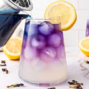 A glass pitcher pours vibrant butterfly pea flower lemonade into an iced glass with a purple hue, garnished with a lemon slice. Two other similar drinks are in the background.