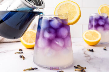 A glass pitcher pours vibrant butterfly pea flower lemonade into an iced glass with a purple hue, garnished with a lemon slice. Two other similar drinks are in the background.