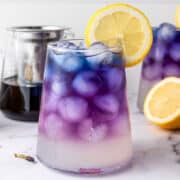 A glass of butterfly pea flower lemonade iced tea boasts an ombre purple to clear gradient, garnished with a lemon slice. Lemons and another glass stand in the background on a marble surface.