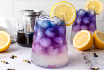 A glass of butterfly pea flower lemonade iced tea boasts an ombre purple to clear gradient, garnished with a lemon slice. Lemons and another glass stand in the background on a marble surface.