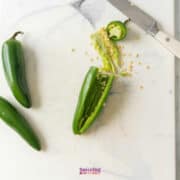 Three jalapeños on a cutting board, one sliced open with seeds exposed and a knife nearby.