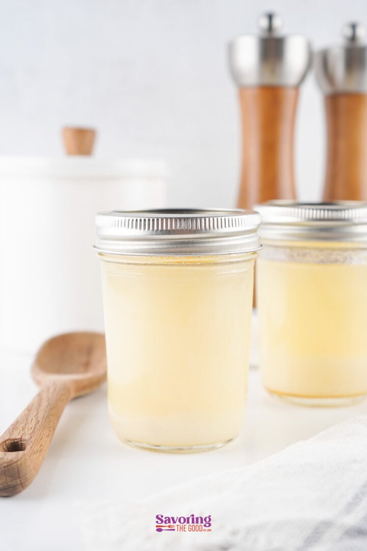Two jars of homemade chicken broth are on a white surface, with a wooden spoon, salt, and pepper grinders in the background.