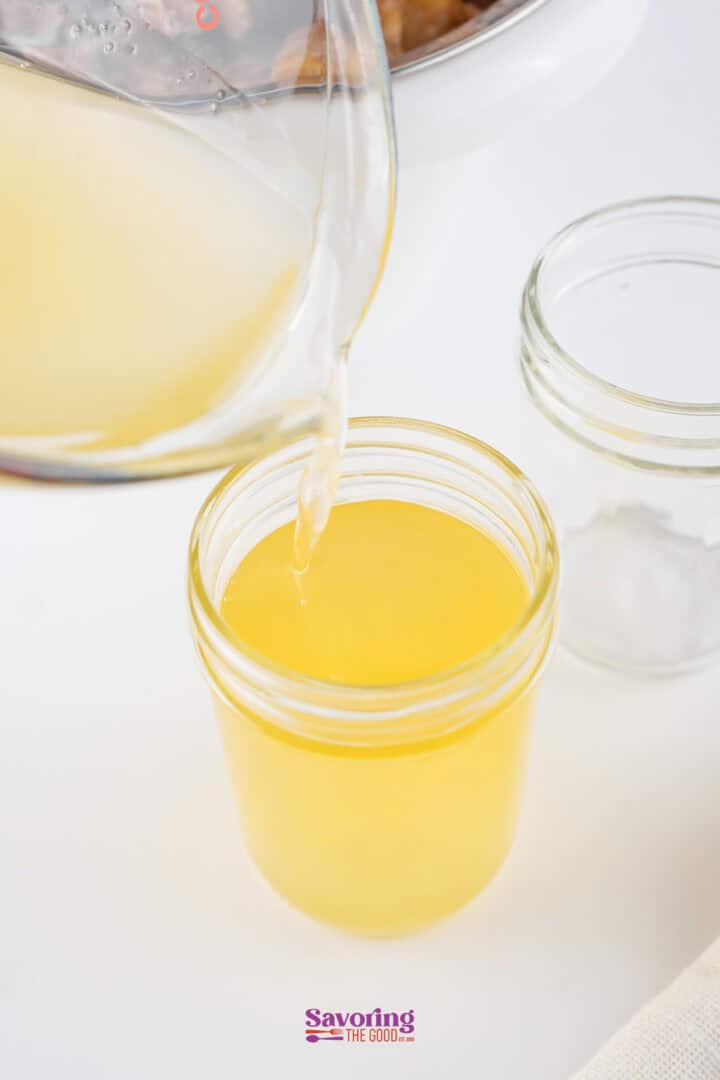 A clear yellow liquid is being poured from a measuring cup into a glass jar on a white surface, with an empty jar and white towel nearby.