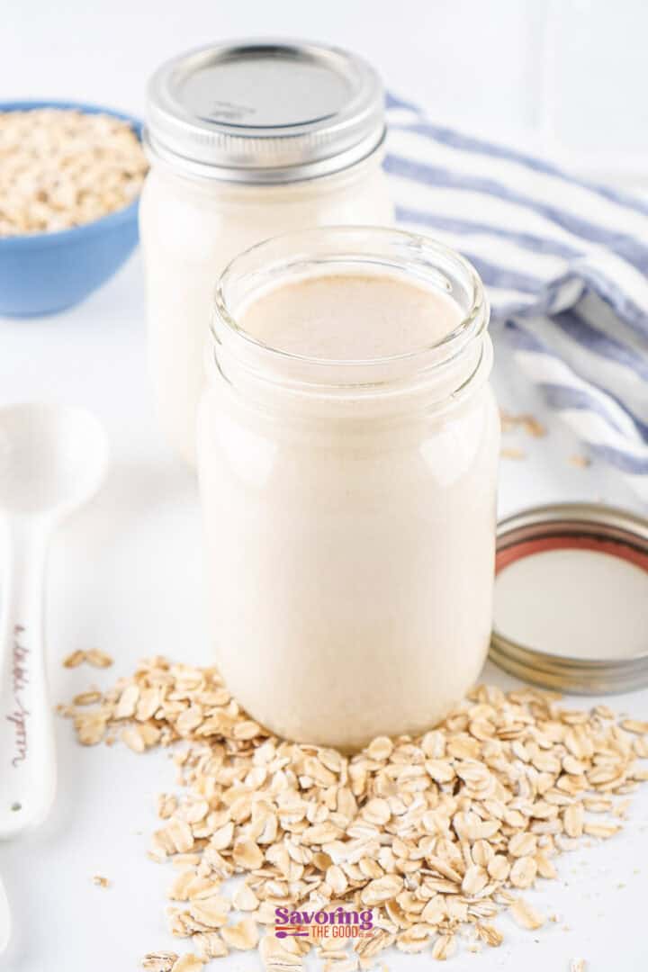 Two glass jars filled with oat milk sit on a white surface, surrounded by scattered oats. A striped cloth and a blue bowl filled with oats are visible in the background.