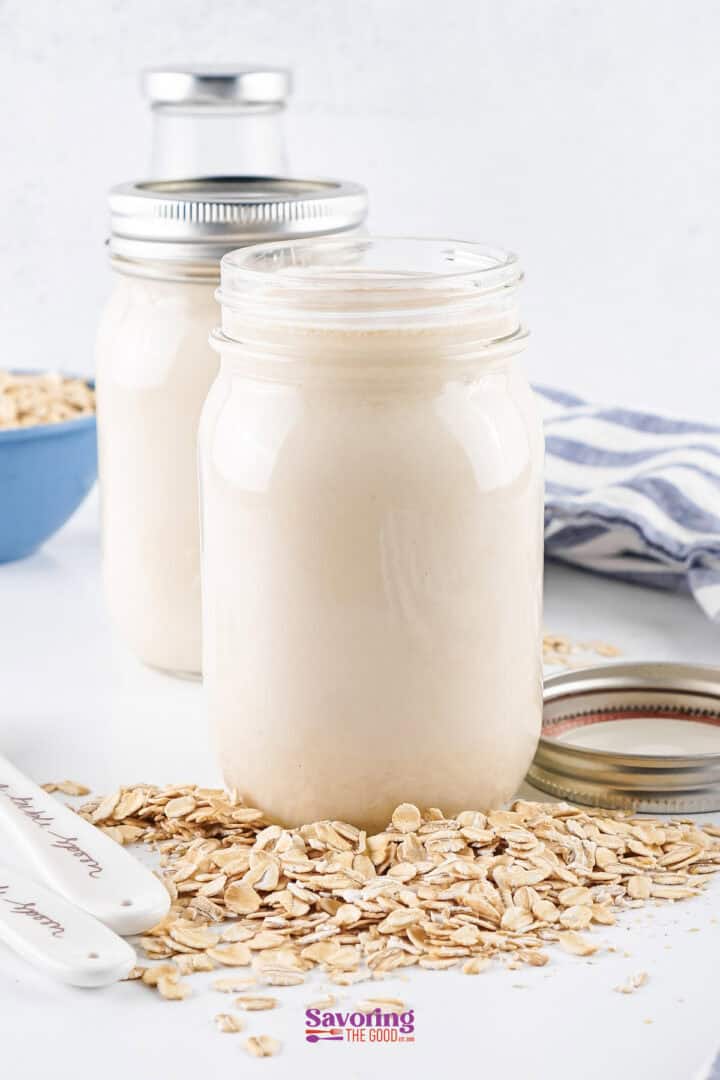 Two jars of oat milk surrounded by rolled oats on a white surface, with a striped cloth and a blue bowl in the background.