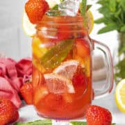 A mason jar filled with iced strawberry lemonade, the perfect mocktail with strawberries, garnished elegantly with lemon slices and mint leaves. A red cloth and whole strawberries add a charming touch beside the jar.