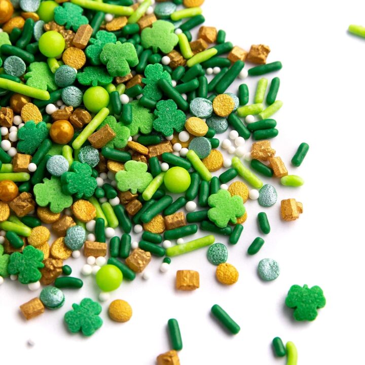 A mix of green, gold, and silver sprinkles, including shamrock shapes, round beads, and rods, scattered on a white surface.