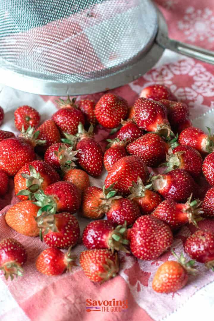 A pile of fresh strawberries on a red and white cloth next to a metal strainer.