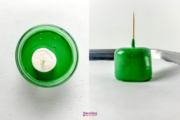 A marshmallow on a skewer is being dipped into green candy coating, shown in a small cup and placed on a parchment-lined tray.
