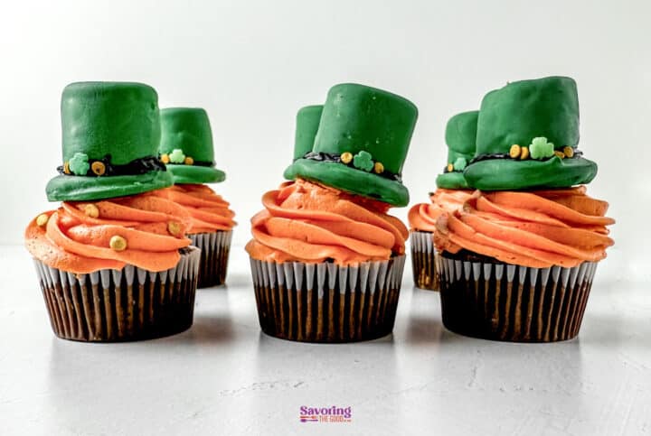 Six Saint Patrick's Day cupcakes with orange frosting are topped with green fondant hats, perfectly resembling leprechaun hats, on a light background.
