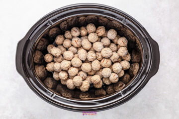 A slow cooker brimming with uncooked, breaded French onion meatballs rests invitingly on a pristine white surface.