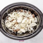 A crockpot brimming with French onion meatballs is layered with sliced onions and topped off with aromatic minced garlic.