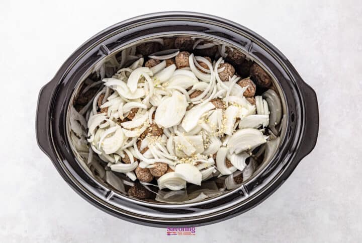 A crockpot brimming with French onion meatballs is layered with sliced onions and topped off with aromatic minced garlic.