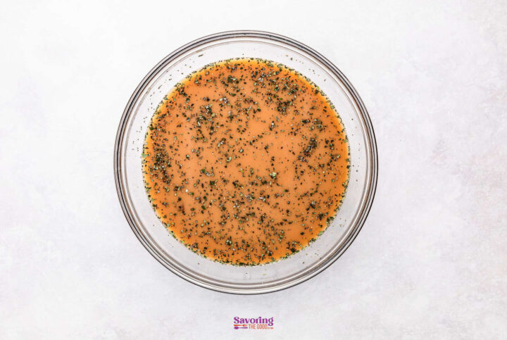 A glass bowl of light brown marinade with herbs, perfect for your French onion meatballs, rests on a plain white surface.