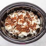 A crockpot brimming with French onion meatballs, sliced onions, and spices rests elegantly on a white surface.