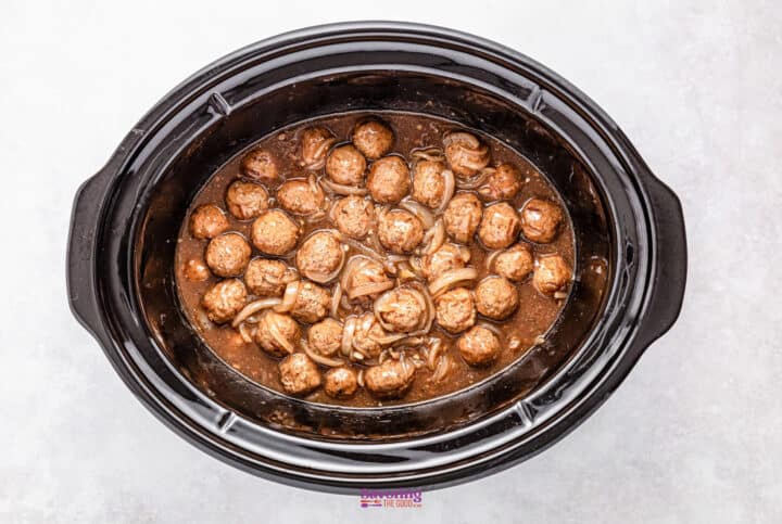 A crockpot brimming with French onion meatballs, bathed in a savory brown sauce and mingling with tender sliced onions.
