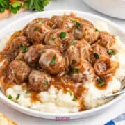 French onion meatballs in gravy rest atop creamy mashed potatoes, garnished with parsley and served in a white bowl with a fork for easy dining.