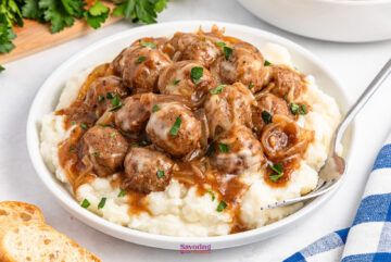 French onion meatballs in gravy rest atop creamy mashed potatoes, garnished with parsley and served in a white bowl with a fork for easy dining.