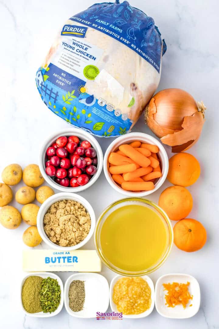 Whole raw chicken with ingredients including onions, baby carrots, cranberries, potatoes, oranges, brown sugar, butter, orange zest, thyme, and olive oil displayed on a marble surface.