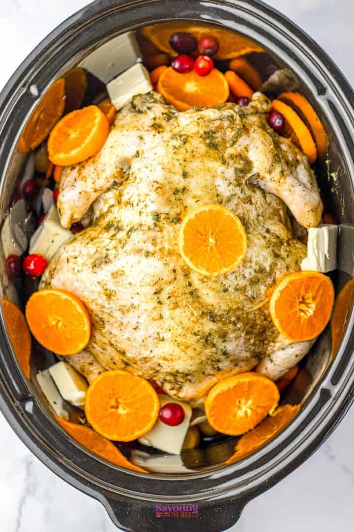 Whole chicken in a slow cooker surrounded by orange slices, cranberries, and butter cubes, seasoned with herbs.