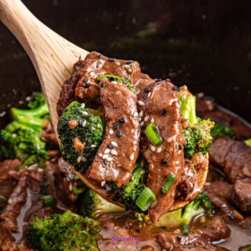 A wooden spoon holds slices of beef with broccoli, garnished with sesame seeds and green onions, in a ceramic bowl.