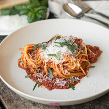 A plate of calabrese pasta with rich tomato sauce, topped with grated cheese and fresh basil. Two spoons and vibrant basil leaves add charm to the background.