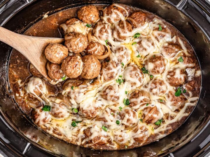 Crockpot meatballs drenched in melted cheese and infused with the rich flavor of French onion soup, a wooden spoon lifting some out.