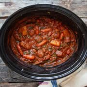 A rustic wooden table holds a slow cooker filled with a savory sausage casserole, brimming with beans, carrots, and potatoes.