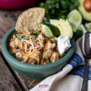 A bowl of McCormick white chicken chili topped with grated cheese, cilantro, avocado slices, lime wedges, a tortilla chip, and a dollop of sour cream sits on a wooden table with a spoon and napkin.