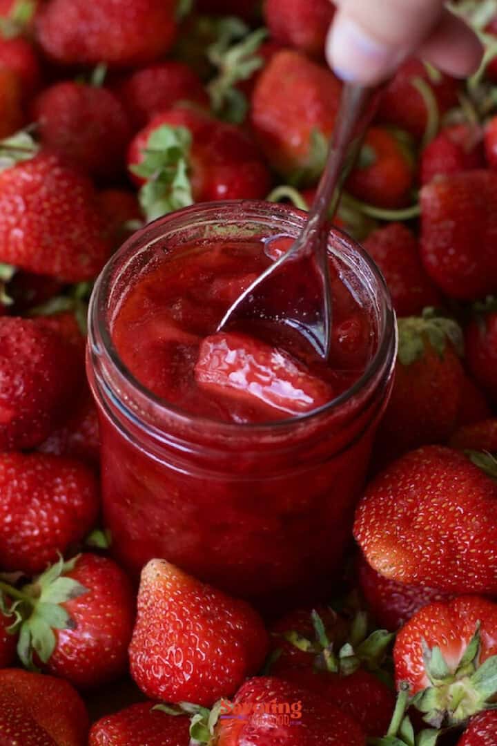 A jar of strawberry jam with a spoon, surrounded by fresh strawberries.