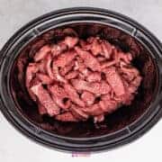 Strips of raw beef and bright green broccoli florets rest inside a black slow cooker on a gray surface, ready to transform into a delicious dish.