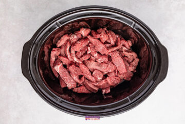 Strips of raw beef and bright green broccoli florets rest inside a black slow cooker on a gray surface, ready to transform into a delicious dish.
