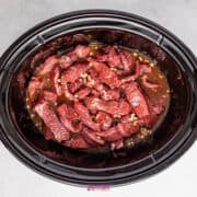 Juicy strips of slow cooker beef marinated in a dark sauce, accompanied by chopped garlic and tender broccoli, simmer delicately together in a black slow cooker on a gray surface.