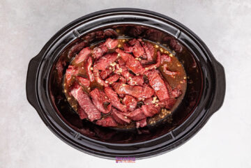 Juicy strips of slow cooker beef marinated in a dark sauce, accompanied by chopped garlic and tender broccoli, simmer delicately together in a black slow cooker on a gray surface.