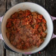 A large pot with a calabrese pasta, featuring diced meat and a rich tomato sauce mixture, rests on a wooden surface.