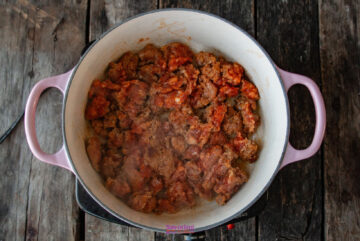 A large pot with a calabrese pasta, featuring diced meat and a rich tomato sauce mixture, rests on a wooden surface.