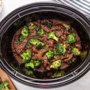 Beef and broccoli in sauce inside a slow cooker, surrounded by cooked rice, chopped scallions, and sesame seeds on a white surface with a striped cloth.