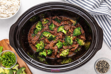 Beef and broccoli in sauce inside a slow cooker, surrounded by cooked rice, chopped scallions, and sesame seeds on a white surface with a striped cloth.
